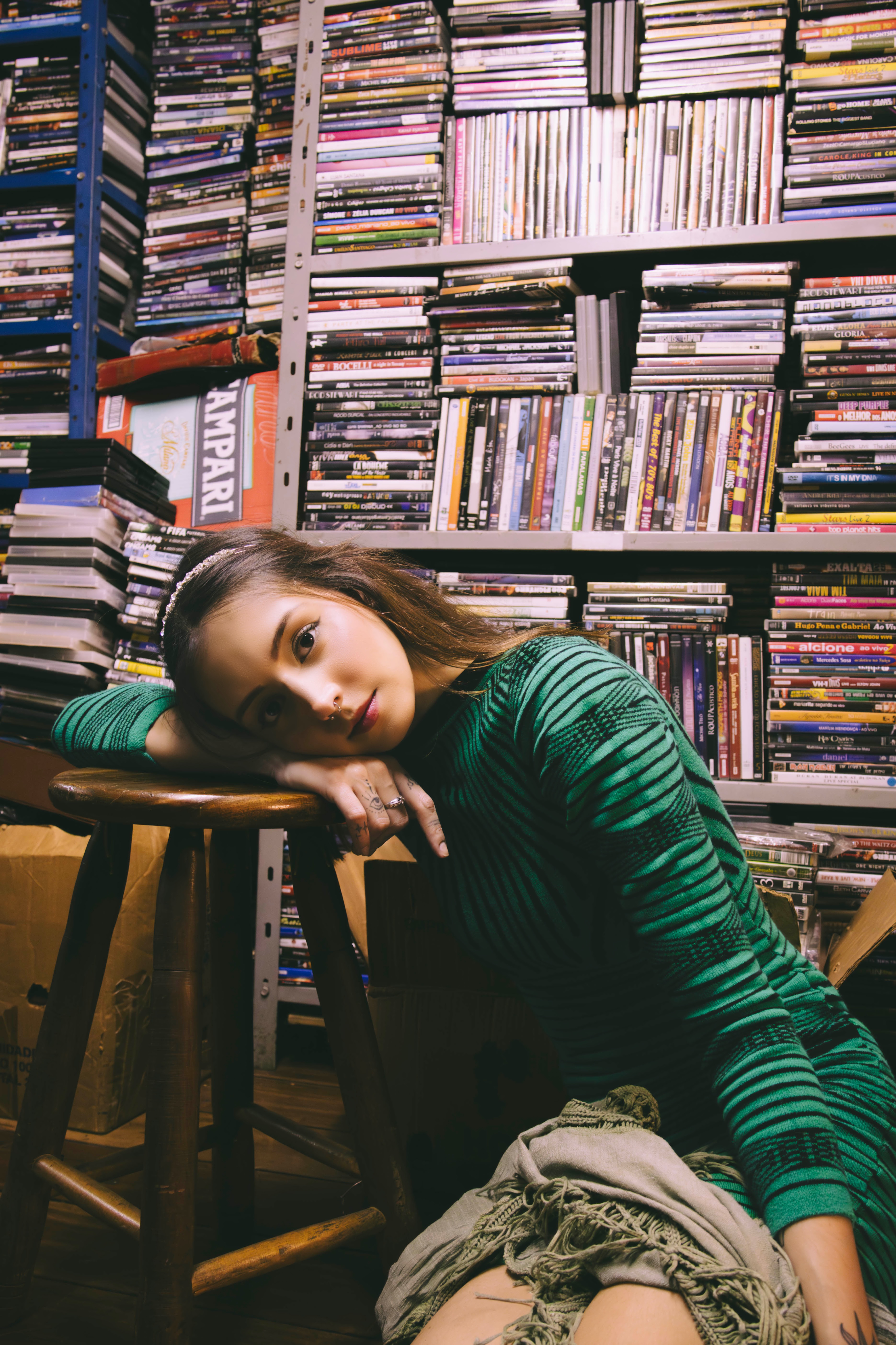 Bárbara Luiza encostada em um banco de madeira, em frente a uma estante repleta de CDs e DVDs